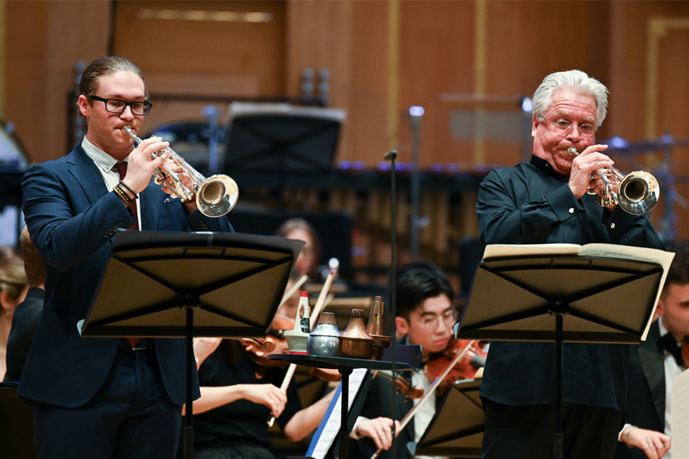 Two trumpet players, both wearing smart black suits and glasses, performing in an orchestra performance, with the orchestra performing behind them.
